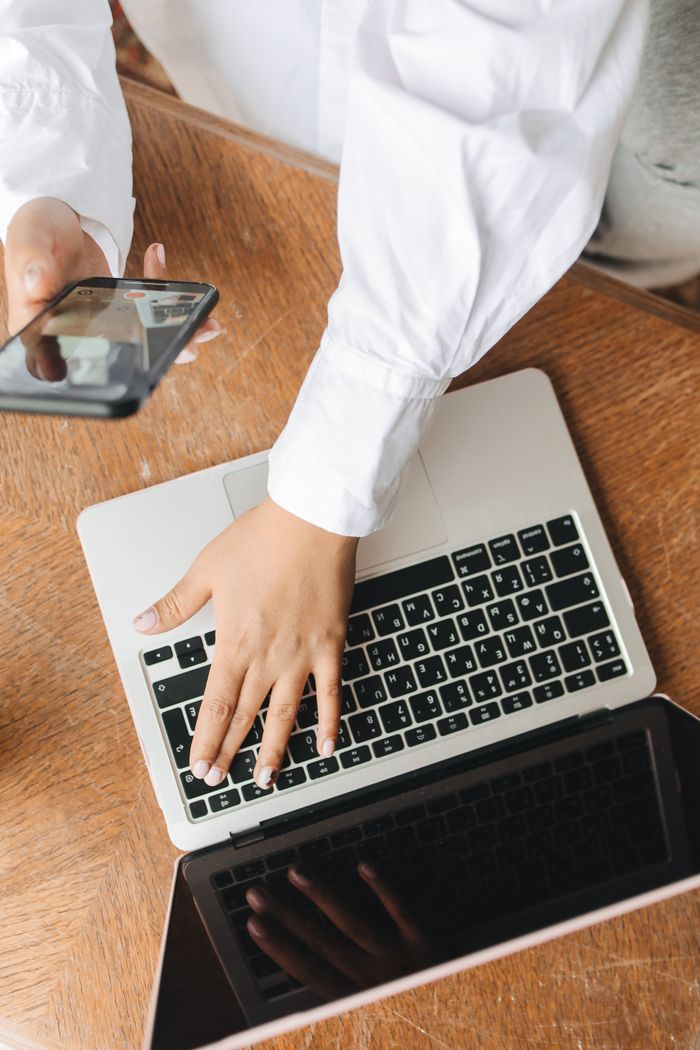Hands typing on a laptop while holding a smartphone, showing a multitasking work environment.